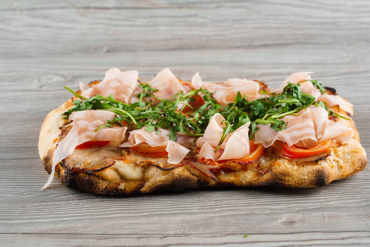 Scrocchiarella gourmet italian cuisine on grey wooden background. Pinsa romana traditional dish. Food delivery from pizzeria. Pinsa with meat, arugula, cheese.